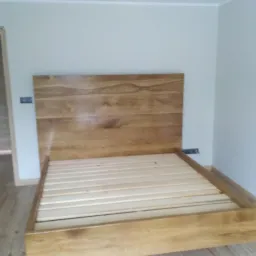 Drewniane łóżko z zagłówkiem z widocznymi słojami drewna, stelaż z listew, surowe ściany z gniazdkiem elektrycznym, drzwi w tle po lewej.