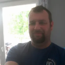 Portret mężczyzny z brodą na tle okna z widokiem na zieleń. Ubrany w niebieską koszulkę z nadrukiem. Uśmiechnięty, naturalne oświetlenie zza okna.