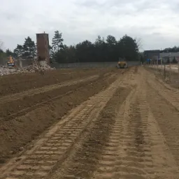 Żółta spycharka wyrównuje teren po rozbiórce budynku; w tle widoczne resztki gruzu, ceglana wieża i drzewa.