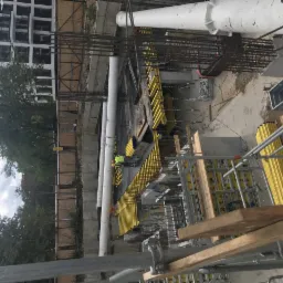 Budowa fundamentów w Warszawie: widok z góry na szalunki, stal zbrojeniową i pracowników w kamizelkach odblaskowych na placu budowy, w tle budynek mieszkalny i drzewa.