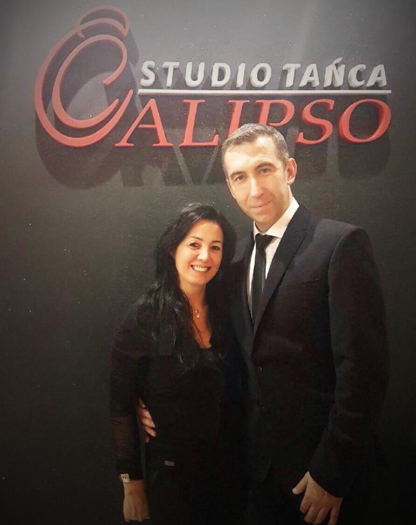 Dwoje uśmiechniętych instruktorów tańca pozuje przed logotypem 'Studio Tańca Calipso' na ciemnej ścianie.