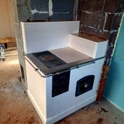 Usługi Zduńskie - Biały piec kaflowy z metalowym obramowaniem płyty grzewczej i drzwiczkami, w trakcie montażu, widoczne surowe ściany i podłoga w remoncie.