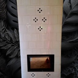 Usługi Zduńskie - Biały, kaflowy kominek z czarnym wkładem, ozdobiony ażurowymi wzorami, na tle ściany w ciemne, roślinne motywy. Widok frontalny, symetryczny układ.