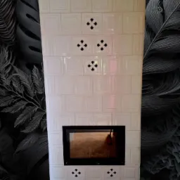 Biały, kaflowy kominek z czarnym wkładem, ozdobiony ażurowymi wzorami, na tle ściany w ciemne, roślinne motywy. Widok frontalny, symetryczny układ.