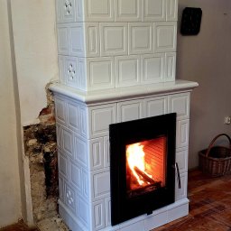 Usługi Zduńskie - Biały, kaflowy kominek z palącym się ogniem, widoczny fragment ściany w trakcie remontu i drewniana podłoga z resztkami po remoncie. Na ścianie wisi zegar.