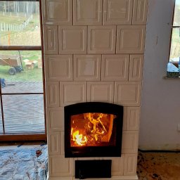 Usługi Zduńskie - Biały, kaflowy kominek z palącym się ogniem, widoczny przez czarną ramę drzwiczek. W tle okno z widokiem na ogród i częściowo zakryta podłoga.