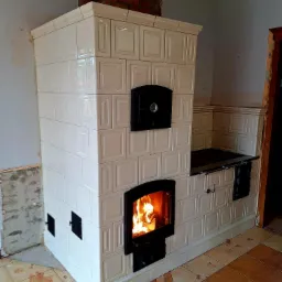 Biały, kaflowy piec z paleniskiem w trakcie rozpalania, widoczne płomienie. Fragment niedokończonej ściany i podłogi w tle. Piec z czarnymi drzwiczkami.