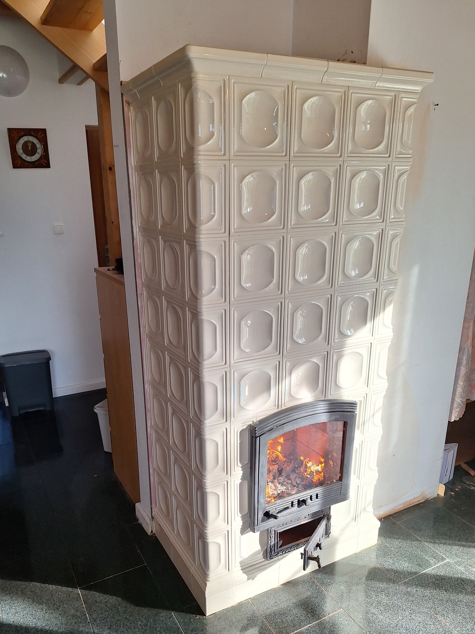 Biały, kaflowy piec z paleniskiem, widoczne płomienie, otwarte drzwiczki popielnika, w tle schody i elementy wyposażenia wnętrza.