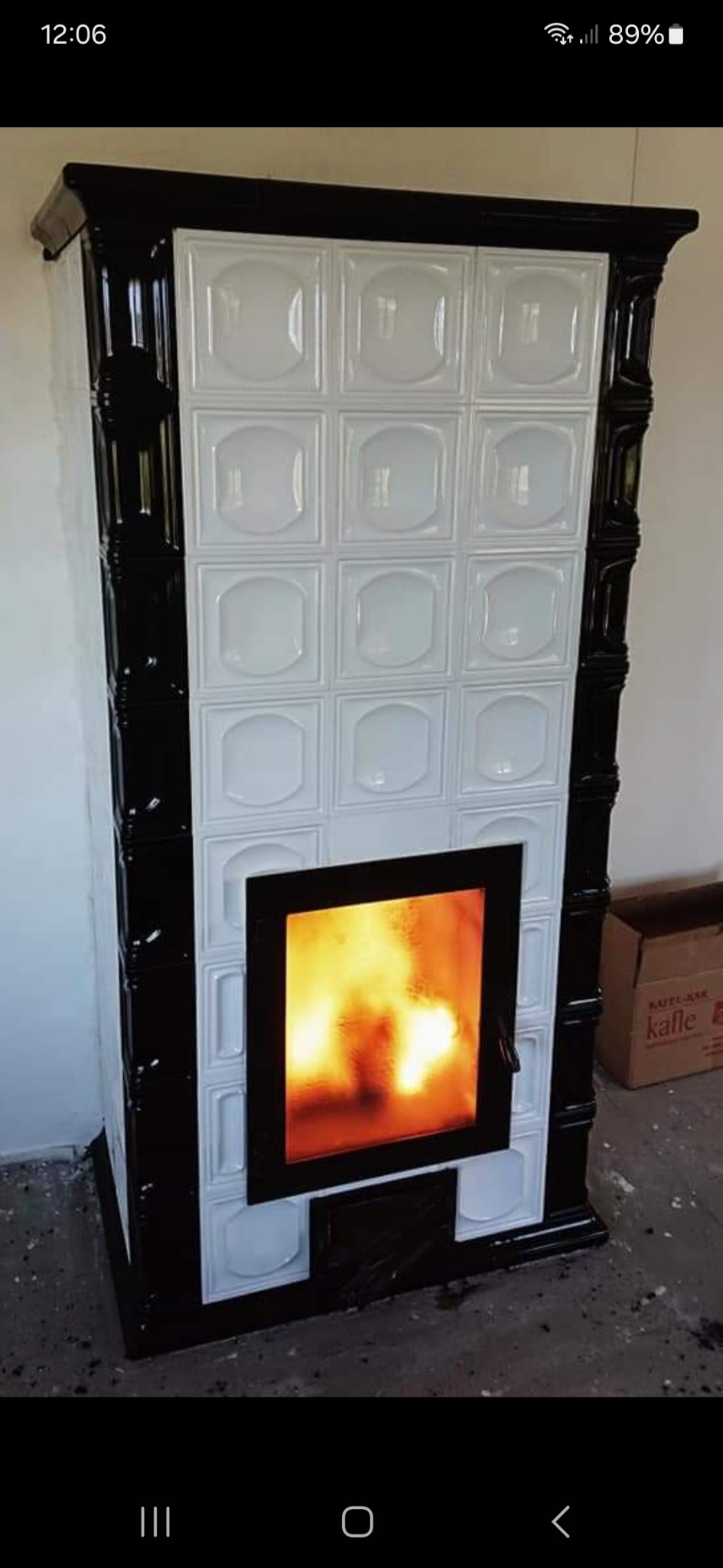 Kafelkowy piec z czarnymi obramowaniami i płomieniami widocznymi przez szybę, ustawiony na surowym podłożu.