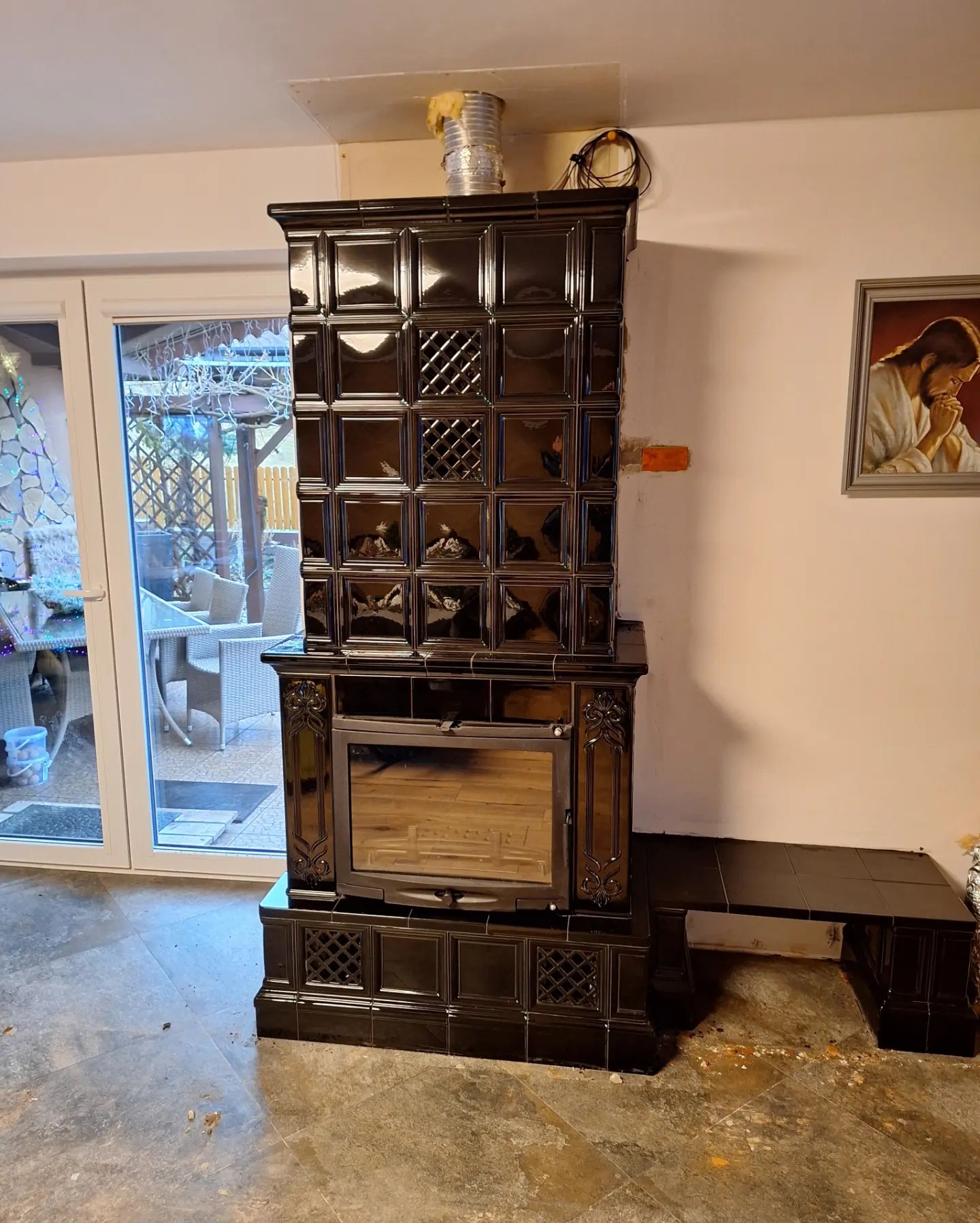 Czarny, kaflowy piec z przeszklonym paleniskiem, widoczne elementy instalacji kominowej, obok okno tarasowe i obraz na ścianie.