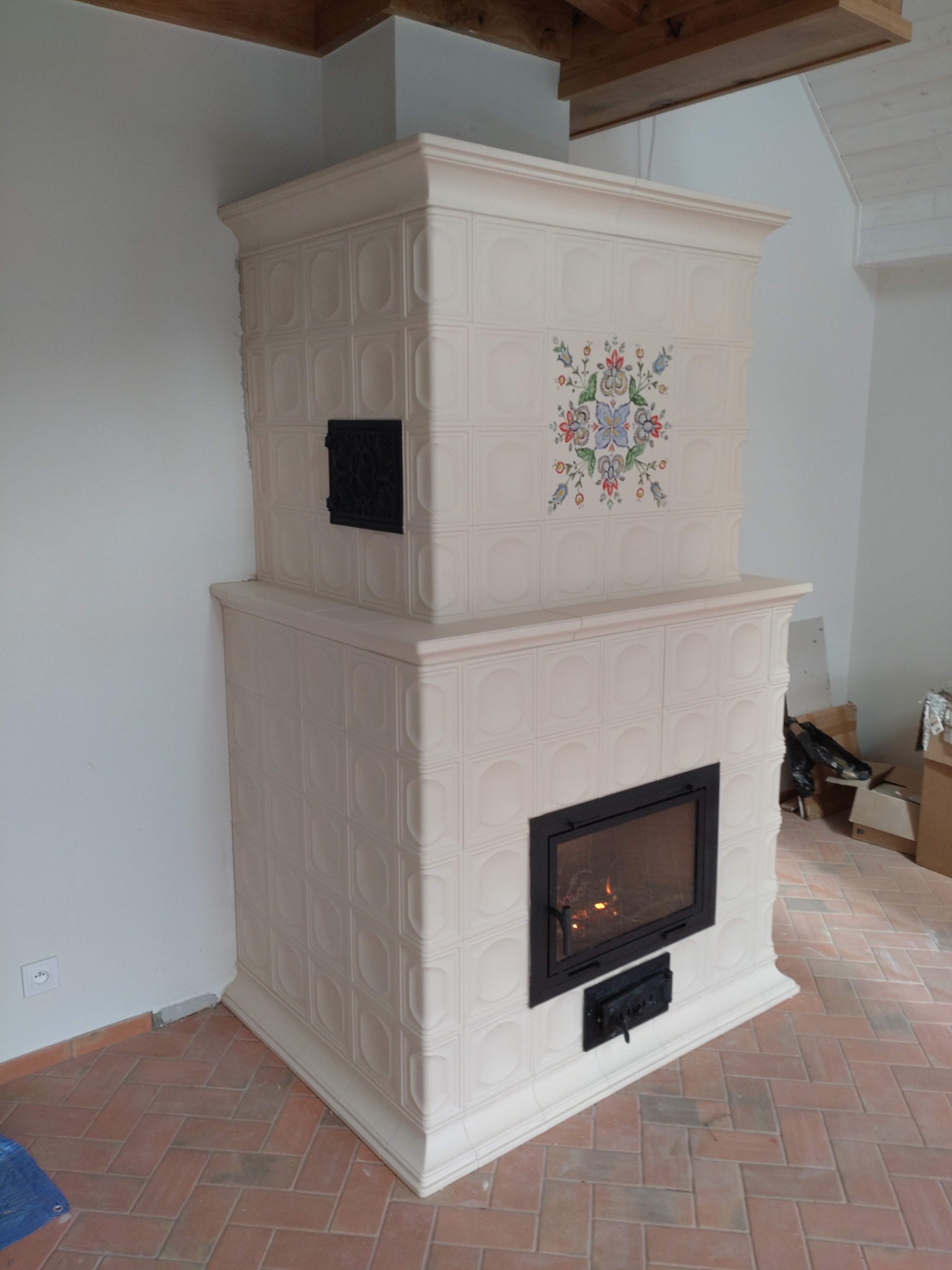 Biały, kaflowy piec z ornamentem kwiatowym i czarnymi drzwiczkami paleniska, ustawiony na ceglanej podłodze w nowo wykończonym pomieszczeniu.