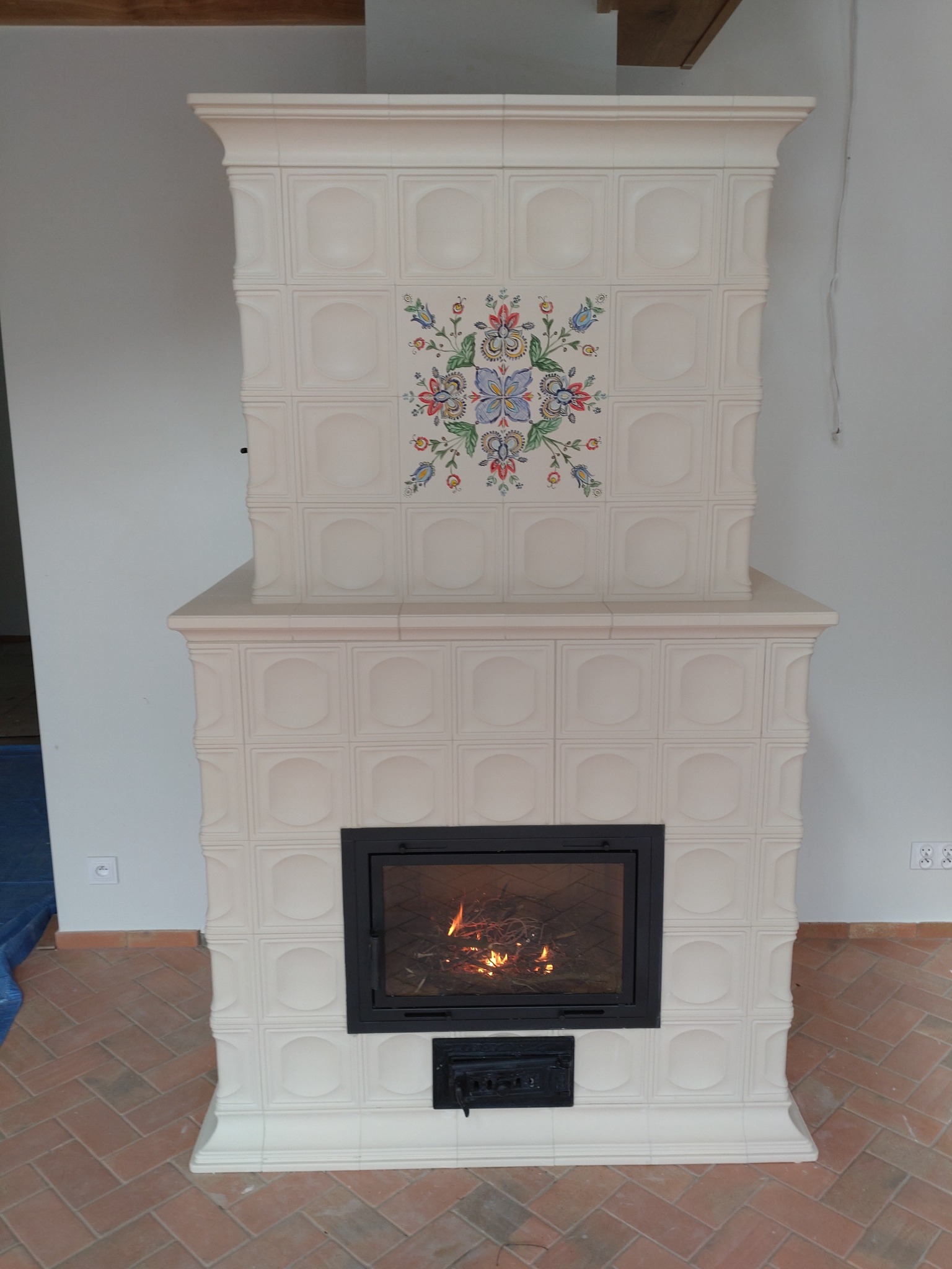 Biały, kaflowy piec z czarną wkładką kominkową, zdobiony folkowym motywem kwiatowym, z palącym się drewnem wewnątrz.