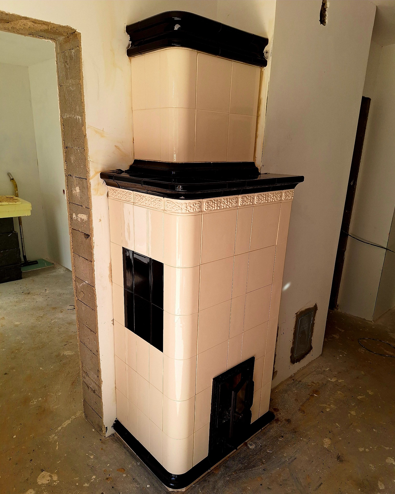 Biały, kaflowy piec z czarnymi akcentami w trakcie montażu, widoczny surowy mur w tle.