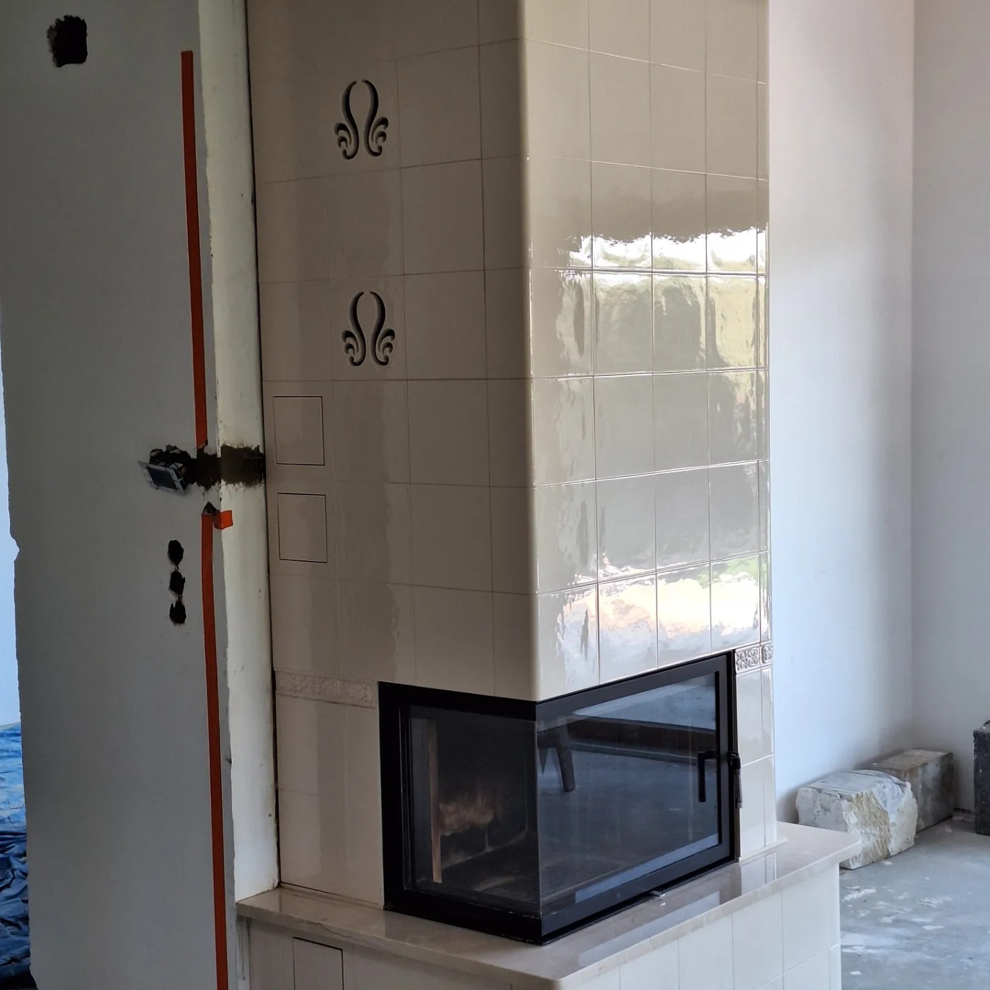 Wnętrze w trakcie wykończenia z widocznym, częściowo obłożonym kremowymi kaflami kominkiem z czarną ramą; na kaflach dekoracyjne motywy, widoczne niedokończone instalacje elektryczne na ścianie.