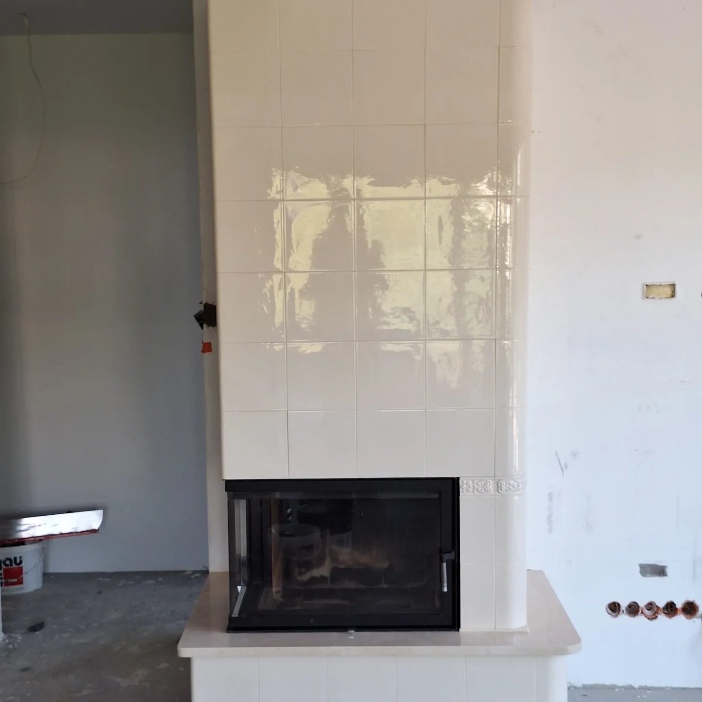 Biały, kafelkowy kominek z czarnym wkładem, w surowym wnętrzu, widoczne niedokończone ściany i narzędzia pracy.