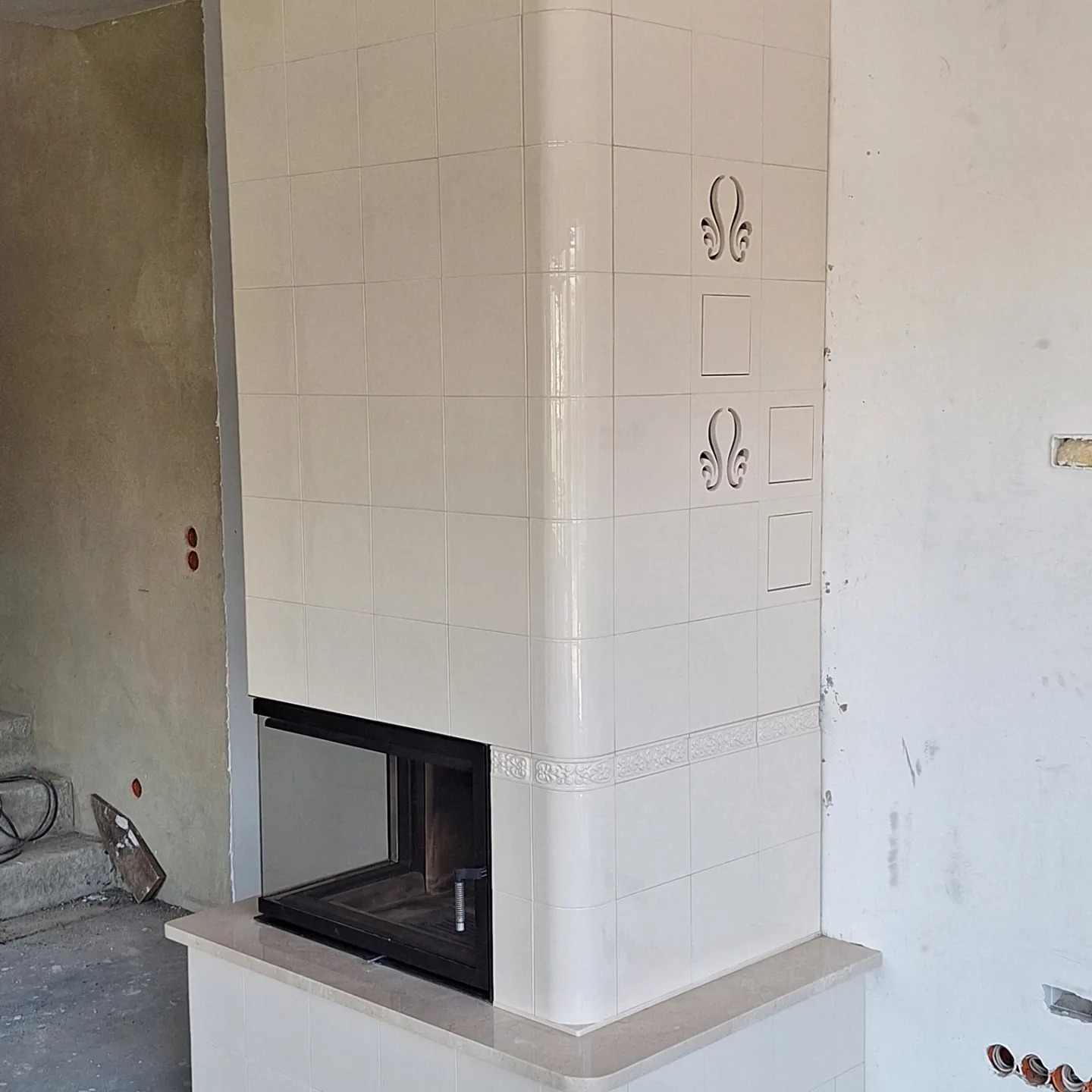 Biały, kaflowy kominek z czarnym wkładem kominkowym w trakcie montażu w Bolszewie, widoczne elementy dekoracyjne i dostęp rewizyjny.