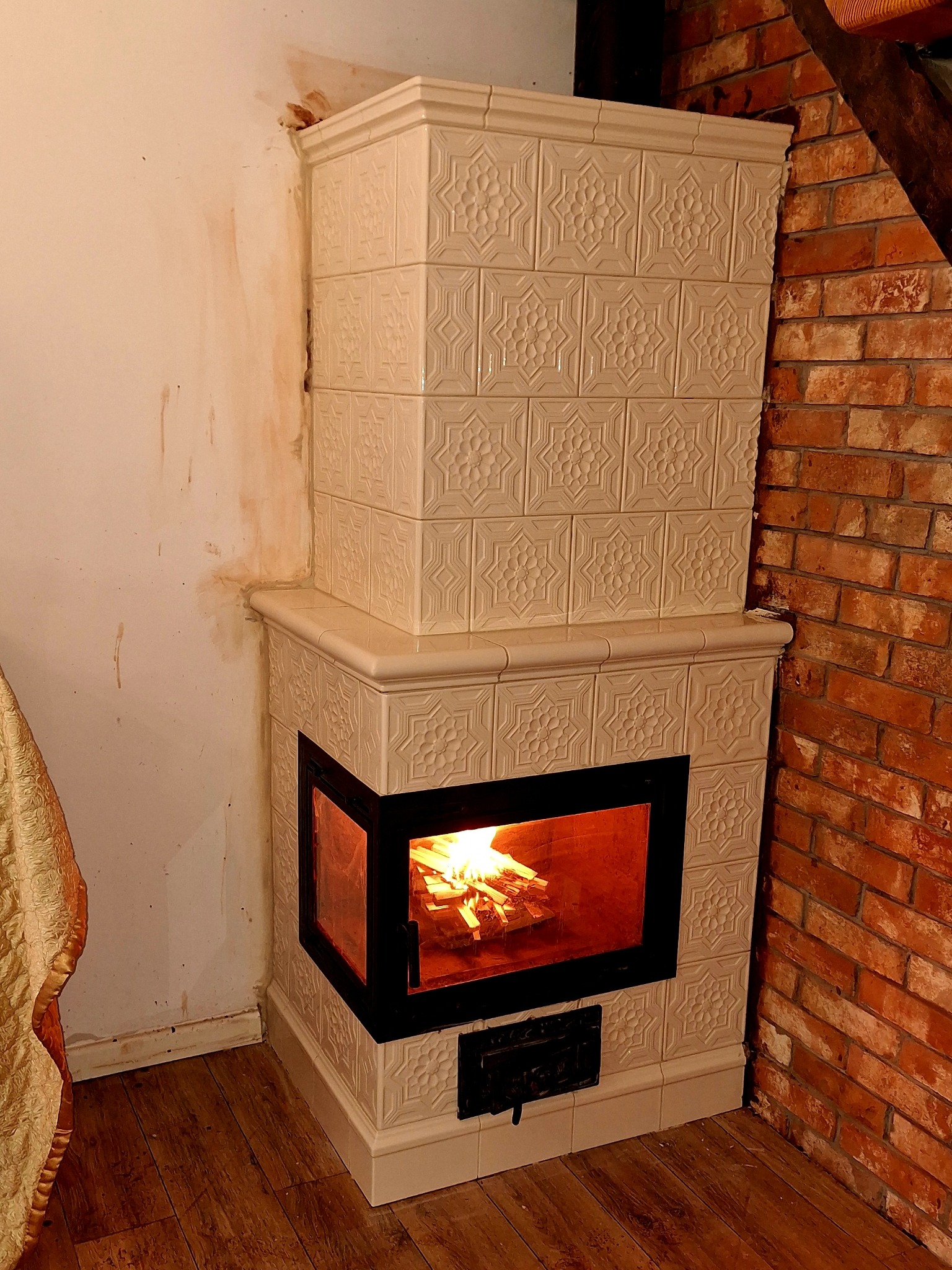 Biały, kaflowy piec kominkowy z paleniskiem, płonące drewno widoczne przez szybę, obok ściana z czerwonej cegły i fragment ściany z widocznymi zabrudzeniami.