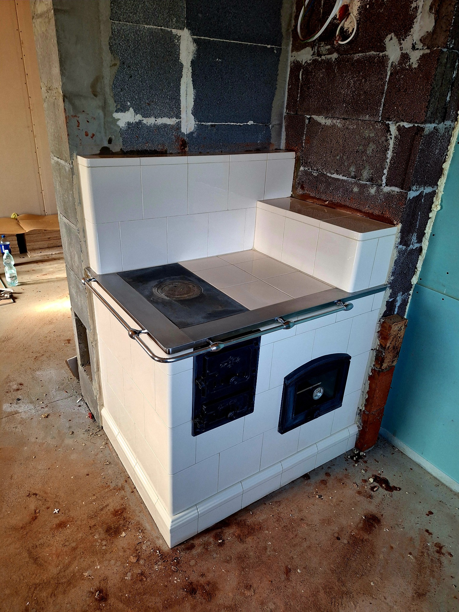 Biały piec kaflowy z metalowym obramowaniem płyty grzewczej i drzwiczkami, w trakcie montażu, widoczne surowe ściany i podłoga w remoncie.