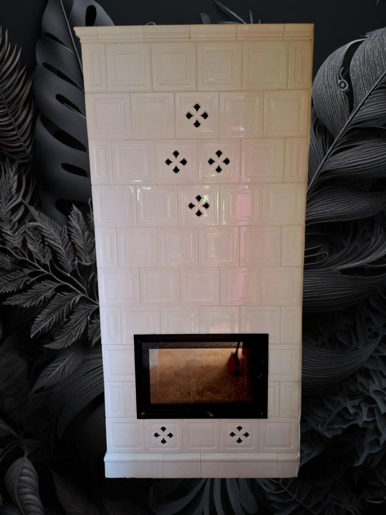 Biały, kaflowy kominek z czarnym wkładem, ozdobiony ażurowymi wzorami, na tle ściany w ciemne, roślinne motywy. Widok frontalny, symetryczny układ.