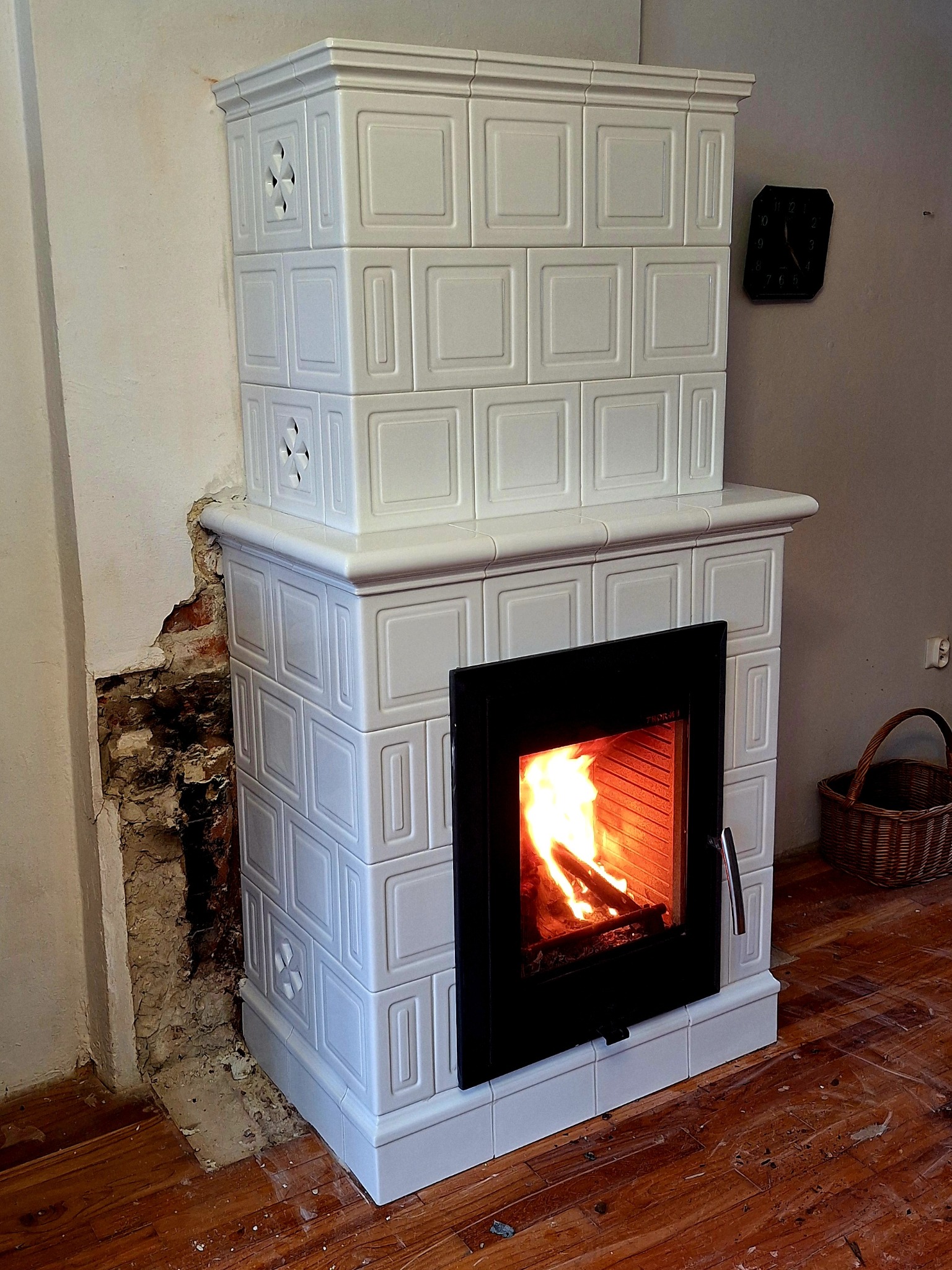 Biały, kaflowy kominek z palącym się ogniem, widoczny fragment ściany w trakcie remontu i drewniana podłoga z resztkami po remoncie. Na ścianie wisi zegar.
