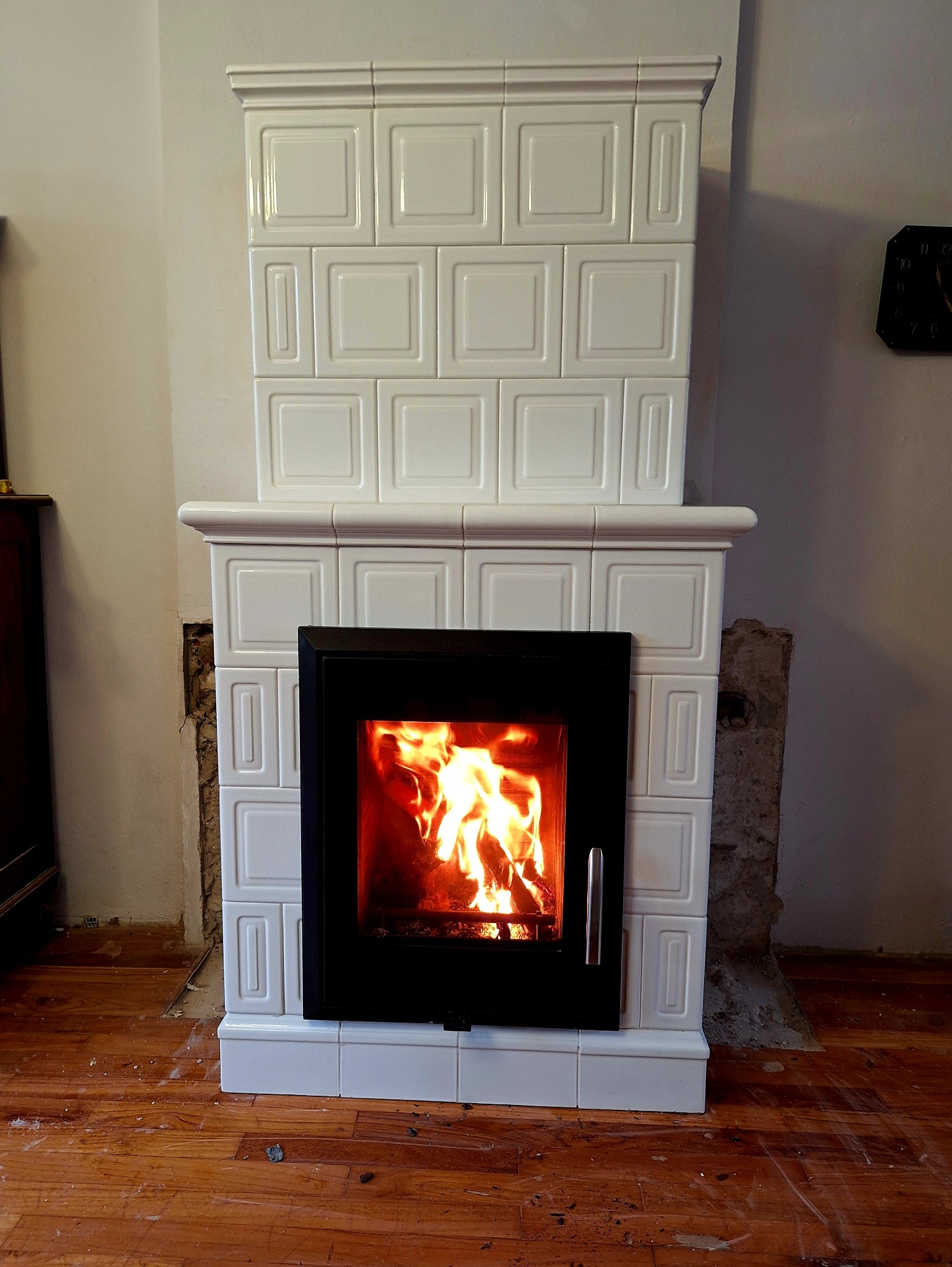 Biały, kaflowy kominek z czarnym wkładem, w którym płonie ogień. Widoczne fragmenty ściany w trakcie prac wykończeniowych i drewniana podłoga.