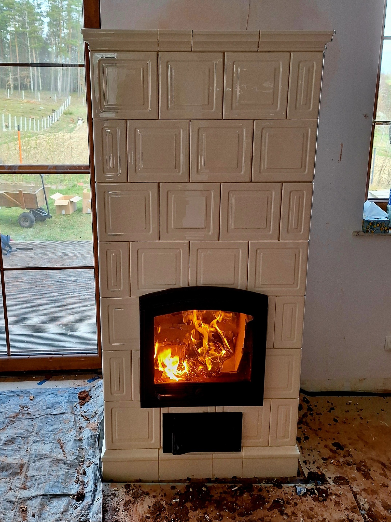 Biały, kaflowy kominek z palącym się ogniem, widoczny przez czarną ramę drzwiczek. W tle okno z widokiem na ogród i częściowo zakryta podłoga.