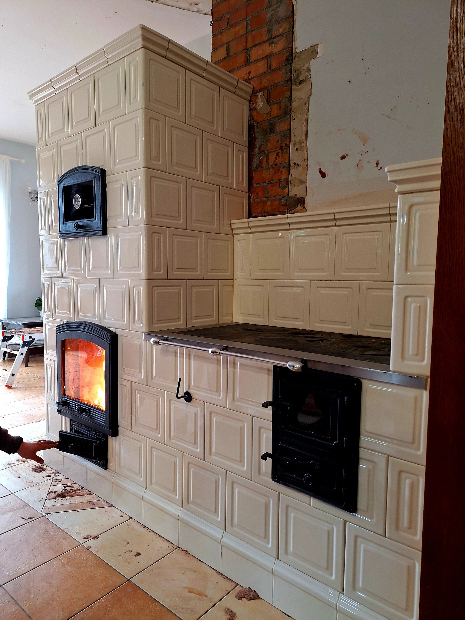 Wykończony piec kaflowy w odcieniach beżu z czarnymi drzwiczkami paleniska i piekarnika, fragment ściany z odsłoniętą cegłą w tle.