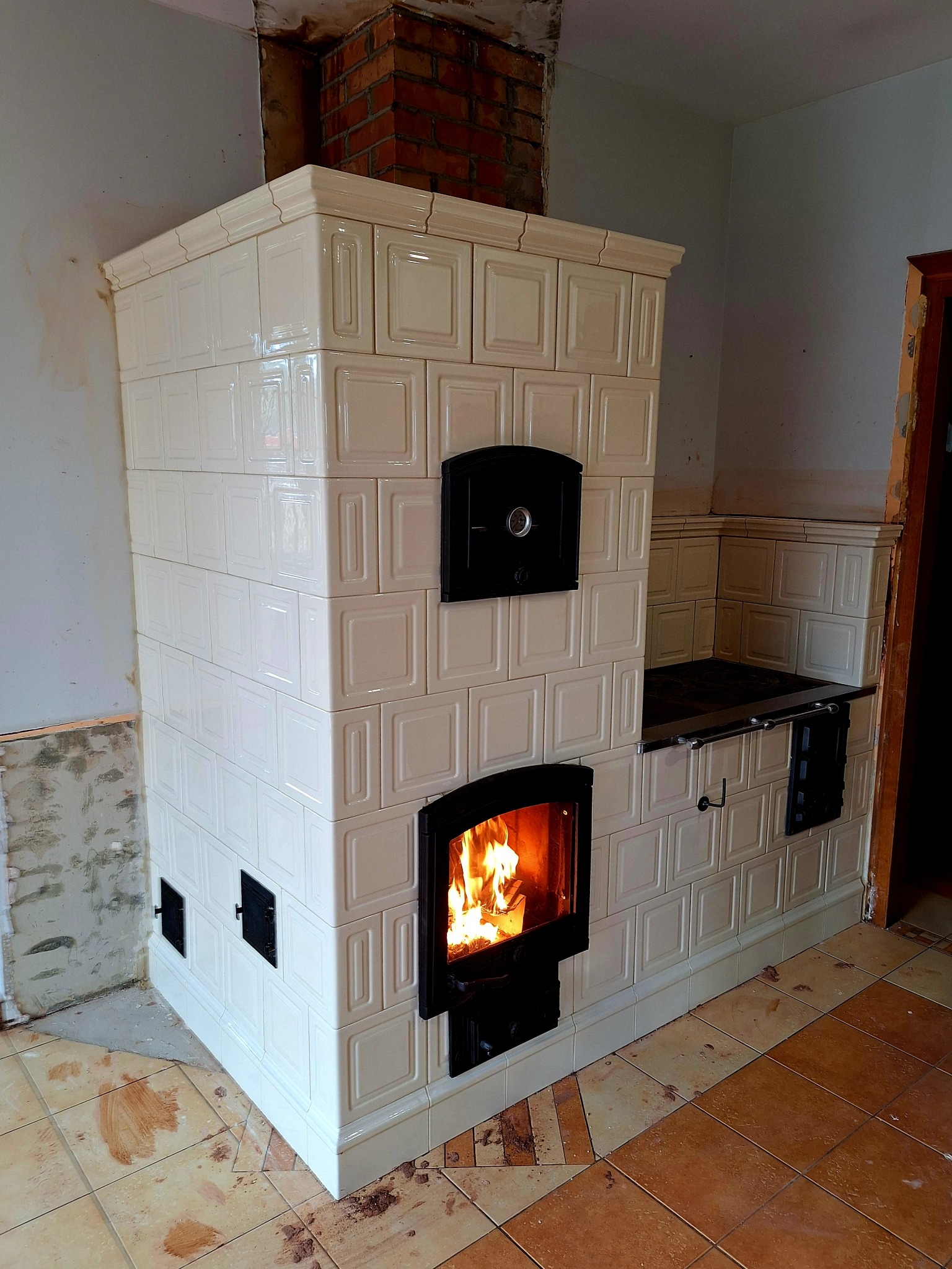 Biały, kaflowy piec z paleniskiem w trakcie rozpalania, widoczne płomienie. Fragment niedokończonej ściany i podłogi w tle. Piec z czarnymi drzwiczkami.