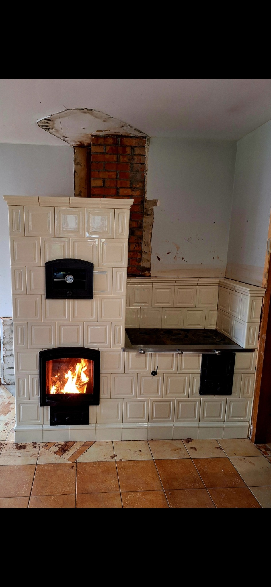 Wykończony piec kaflowy z paleniskiem, widoczne płomienie. Fragment ściany z odsłoniętą cegłą. Beżowe płytki podłogowe. Ukończona realizacja zduna.