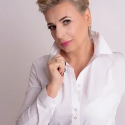 Portret kobiety z krótkimi blond włosami, w białej koszuli z szerokim kołnierzem, z pierścionkiem na palcu, patrzącej w obiektyw, z wyraźnym makijażem oczu i różowymi ustami.