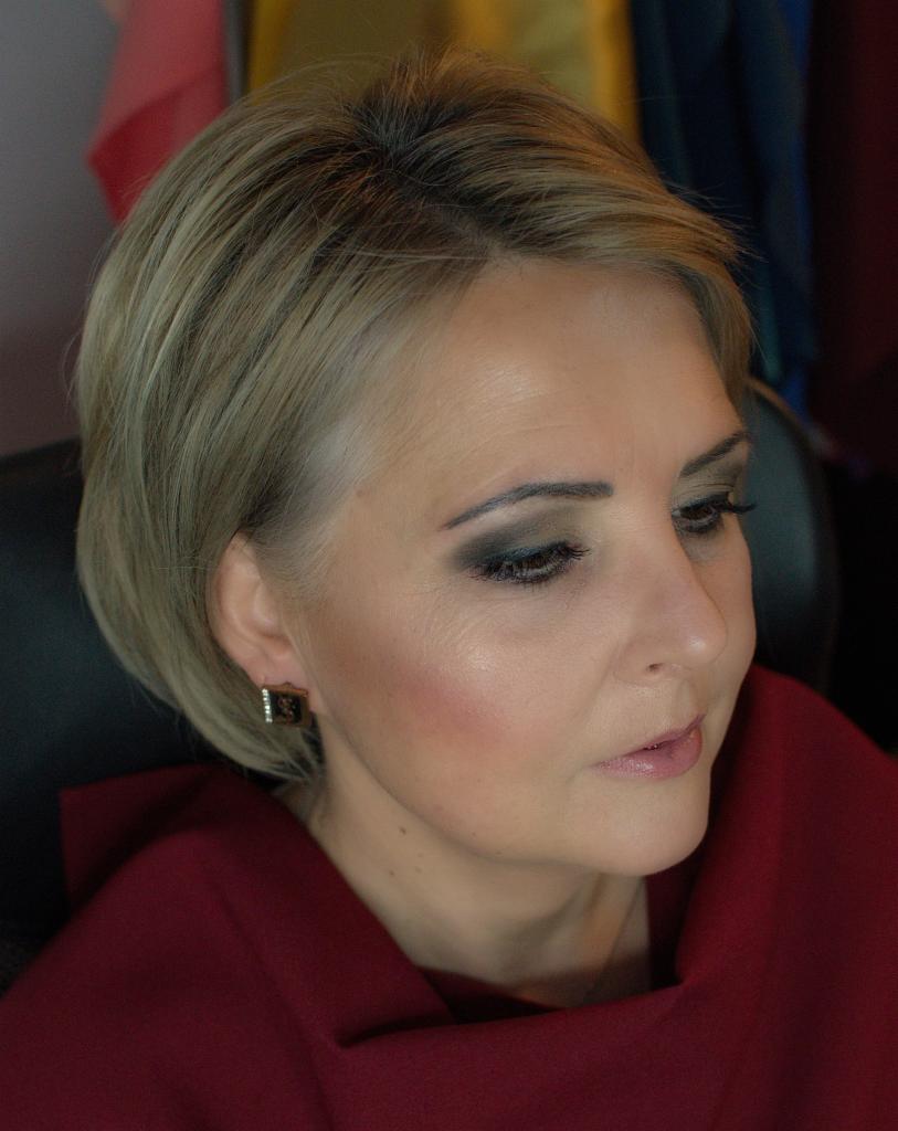 Portret kobiety z blond włosami i wieczorowym makijażem oczu w odcieniach szarości, widoczny kolczyk z błyszczącymi kamieniami i szminka w naturalnym odcieniu.