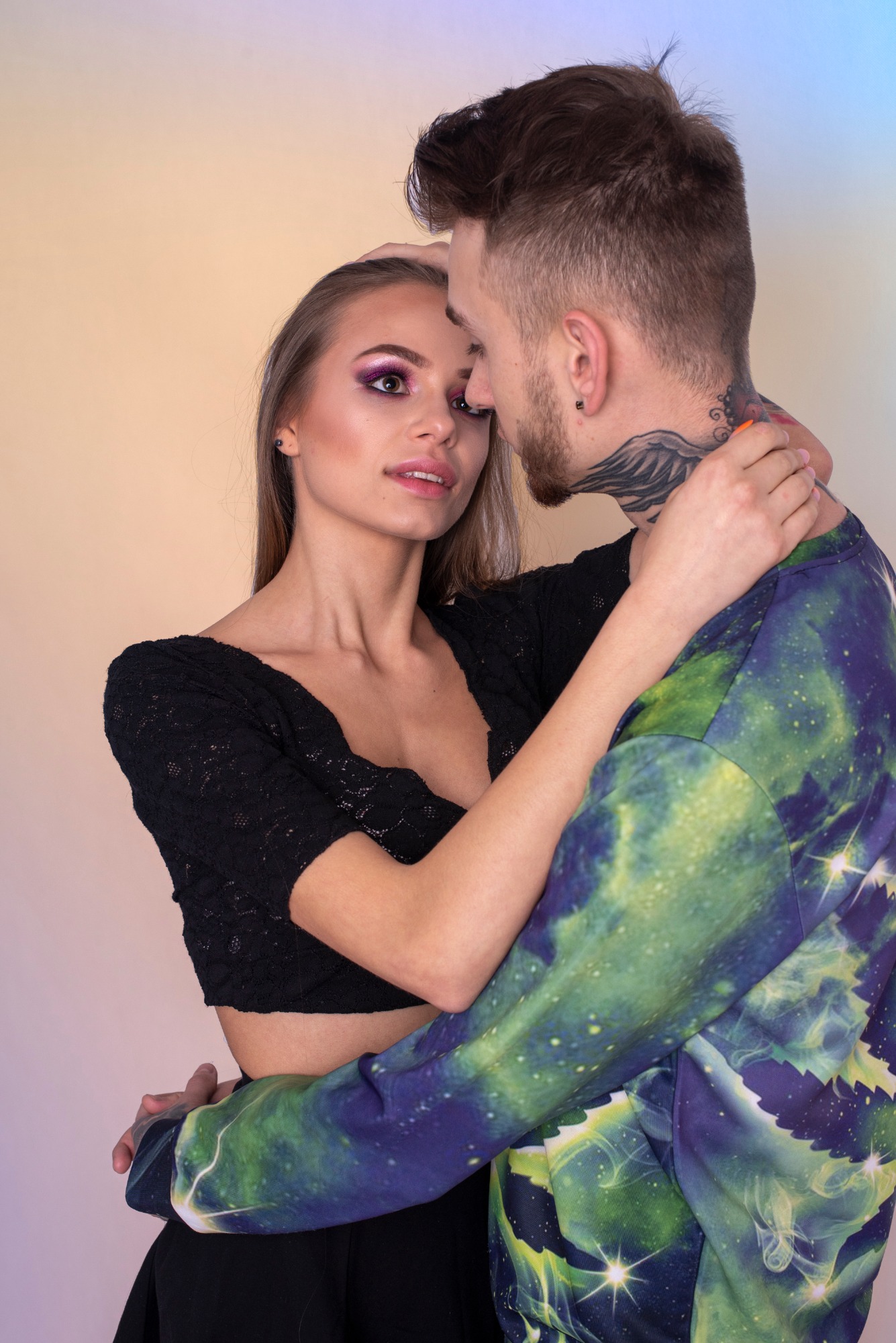 Para obejmująca się na neutralnym tle; kobieta z wyrazistym fioletowym makijażem oczu i czarnej koronkowej bluzce, mężczyzna w bluzie z motywem kosmosu i tatuażem na szyi.