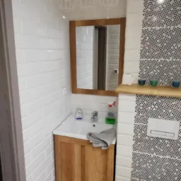 Wykończona łazienka z umywalką w drewnianej szafce, białymi kafelkami typu metro i dekoracyjną ścianą z wzorzystych płytek. Nad umywalką lustro w drewnianej ramie i trzy wiszące lampy z drucianymi...