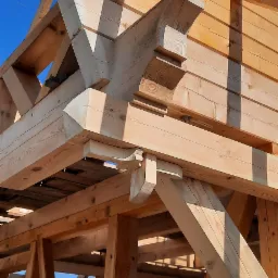 Konstrukcja drewniana domu w trakcie budowy, widoczne elementy z jasnego drewna i złączenia belek, na tle błękitnego nieba.