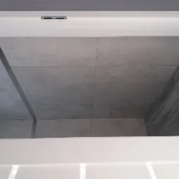 Szare płytki wielkoformatowe z metalową listwą dekoracyjną, ułożone w kabinie prysznicowej, widoczne spod białego sufitu podwieszanego.