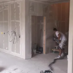 Wnętrze w trakcie prac wykończeniowych: szlifowanie ściany szlifierką z odkurzaczem, widoczne płyty gipsowo-kartonowe na ścianach i suficie, instalacja elektryczna.