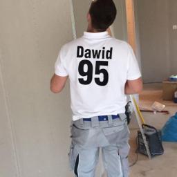 Piotr Słowik - Mężczyzna w roboczym stroju z napisem 'Dawid 95' na plecach, stojący w pomieszczeniu z płytami gipsowo-kartonowymi, montuje narożnik ochronny na ścianie. W tle widoczne narzędzia i materiały...