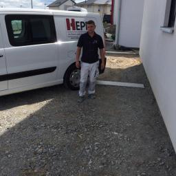 Piotr Słowik - Pracownik w ubraniu roboczym stoi przed białym busem z logo firmy na żwirowym podłożu, obok świeżo pomalowanej ściany budynku.