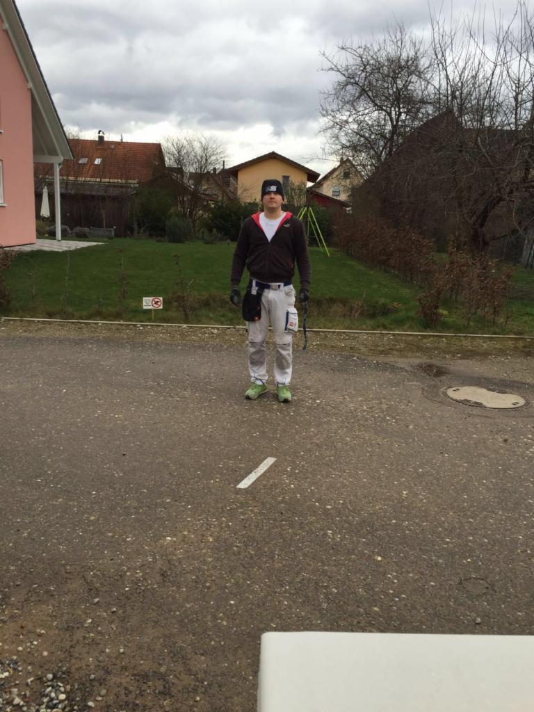 Mężczyzna w ubraniu roboczym, z pasem narzędzi, stojący na tle domów i trawnika, prawdopodobnie malarz przed rozpoczęciem pracy.