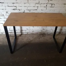 Stół z blatem imitującym drewno i czarnymi metalowymi nogami w kształcie litery V, na tle białej, ceglanej ściany.