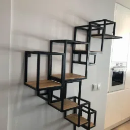 Asymetryczna, geometryczna półka ścienna z czarnych metalowych ram i drewnianych blatów, zamontowana na białej ścianie w nowoczesnym wnętrzu.