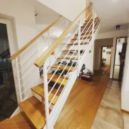 Jasne schody z drewnianymi stopniami i białą, metalową konstrukcją z drewnianą poręczą, widok z dołu na górę w nowoczesnym wnętrzu.