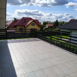 Balkon z czarną, poziomą balustradą z metalu, wyłożony jasnymi płytkami, z widokiem na domy i błękitne niebo z chmurami.