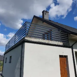 Nowoczesna, pozioma, metalowa balustrada balkonowa w kolorze grafitowym, zamontowana na płaskim dachu budynku z białą elewacją, widok z dołu na tle błękitnego nieba z chmurami.