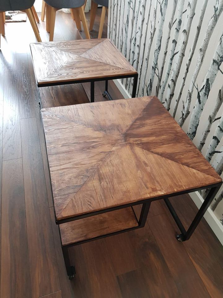 Dwa kwadratowe stoliki kawowe z blatem z drewna o wzorze w jodełkę i czarną metalową ramą, jeden z dodatkową półką, ustawione na drewnianej podłodze przy ścianie z tapetą w brzozy.