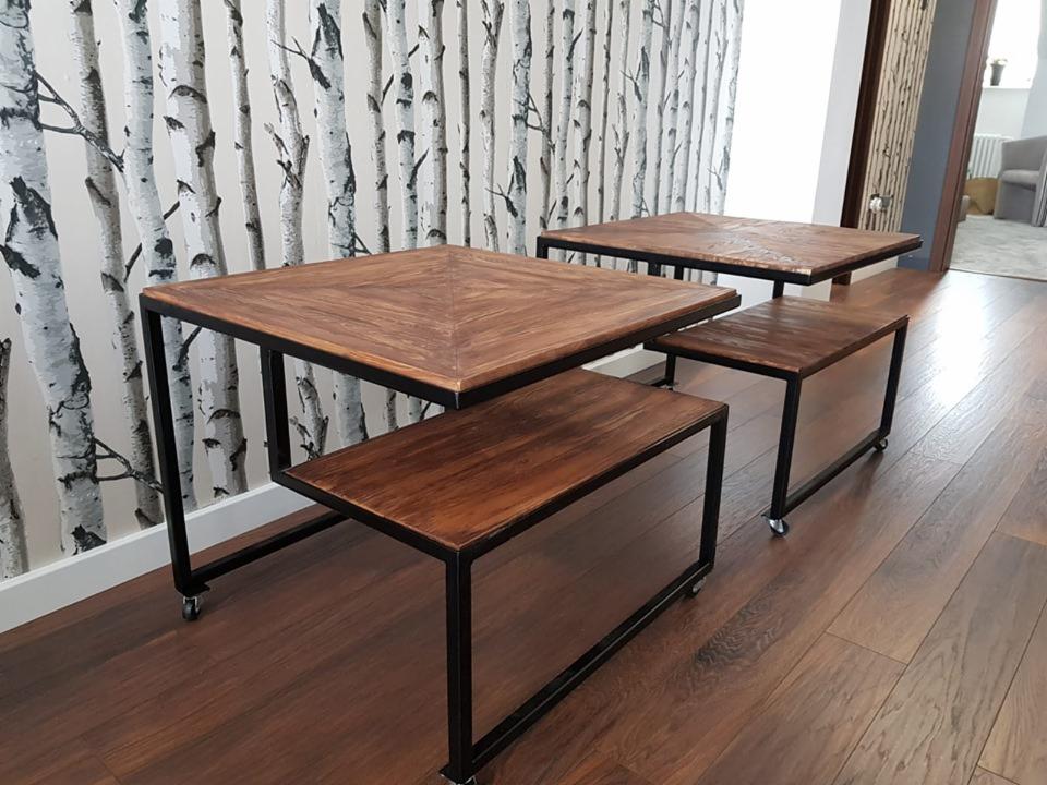 Zestaw dwóch drewnianych stolików i dwóch taboretów z czarnymi metalowymi ramami na kółkach, na tle ściany z tapetą imitującą brzozowy las i podłogi z paneli w odcieniach brązu.