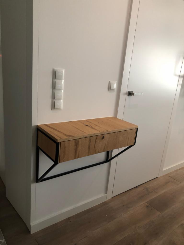 Drewniana szafka nocna z szufladą, wsparta na czarnych, metalowych wspornikach, zamontowana na białej ścianie obok białych drzwi.