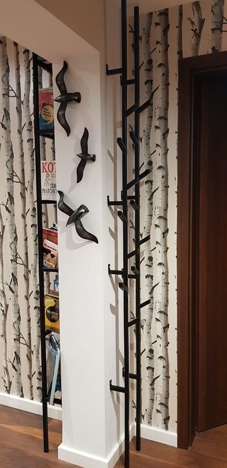 Metalowa konstrukcja przypominająca drzewo z wieszakami, obok regał z książkami i dekoracyjne ptaki na ścianie z tapetą w brzozy, widoczna drewniana podłoga i drzwi.