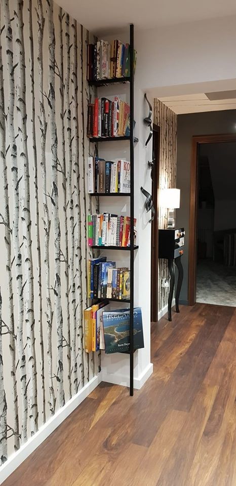 Wąska, czarna metalowa półka na książki, zamontowana w rogu pokoju z tapetą w brzozy, na półkach ułożone książki.