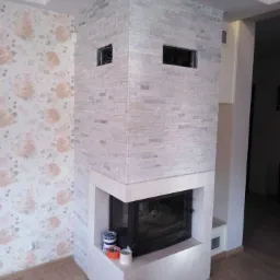 Nowoczesny kominek obłożony jasną, imitującą kamień płytką, z dwoma małymi otworami wentylacyjnymi w górnej części, stoi w rogu pokoju z kwiecistą tapetą i drewnianą podłogą.